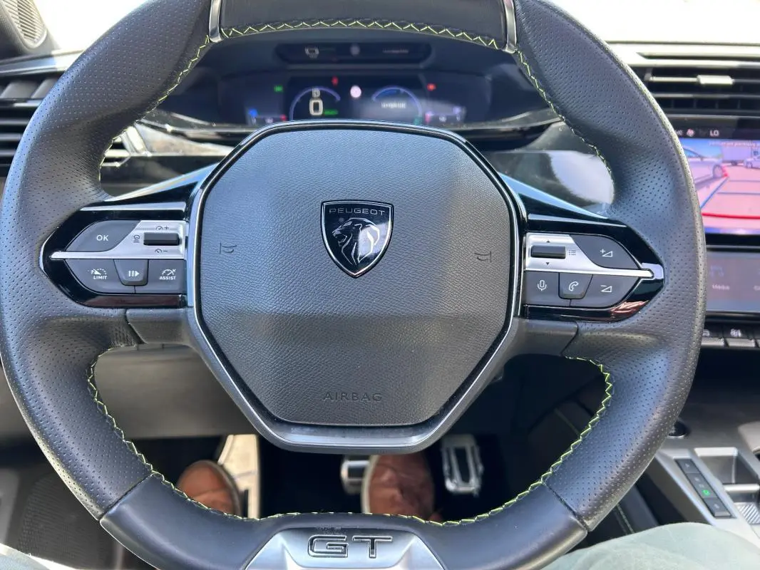 Vue rapprochée du volant cuir noir avec logo Peugeot et badge GT, tableau de bord numérique en arrière-plan.