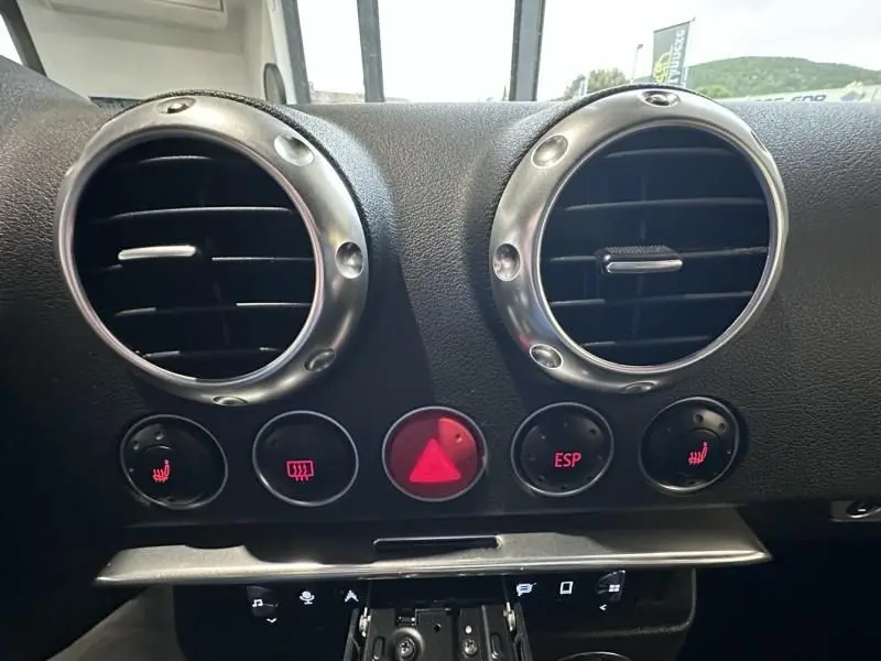 Vue rapprochée des aérateurs ronds chromés et des boutons de commande rouges du tableau de bord d'un Audi TT Coupé noir de 2002.