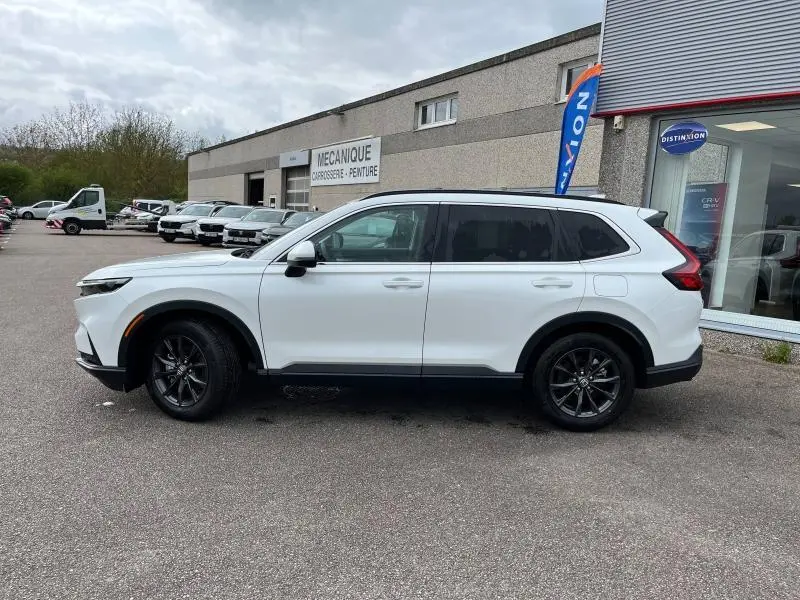 Vue de profil côté gauche d'un Honda CR-V blanc platine nacré 2024 avec jantes alliage noires, stationné devant un garage.