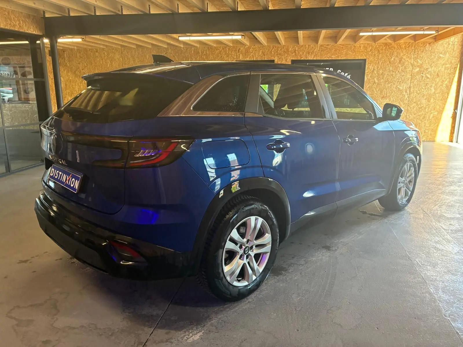 Vue 3/4 arrière droite d’un Renault Austral bleu 2024 dans un garage avec toit en bois, mettant en valeur ses feux arrière et jantes.