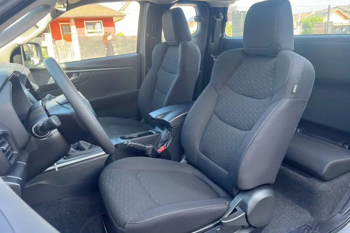 Vue intérieure côté gauche du poste de conduite de l'Isuzu D-MAX 2026 avec sièges tissu gris foncé et accoudoir central.