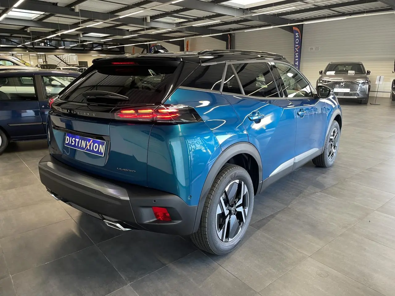 Peugeot 2008 bleu vue 3/4 arrière droit en intérieur avec toit noir et feux arrière LED allumés.