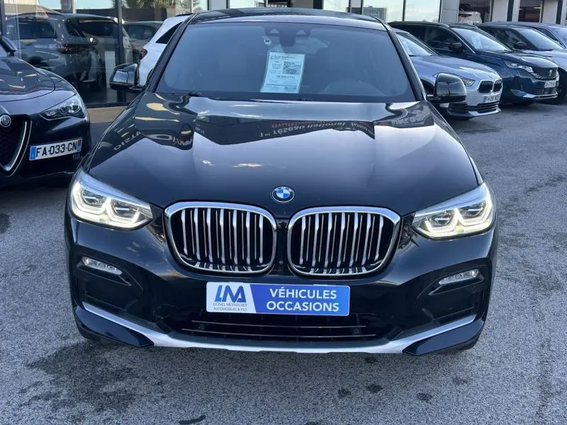 BMW X4 X Drive gris foncé vue de face avec calandre imposante et phares allumés sur parking extérieur