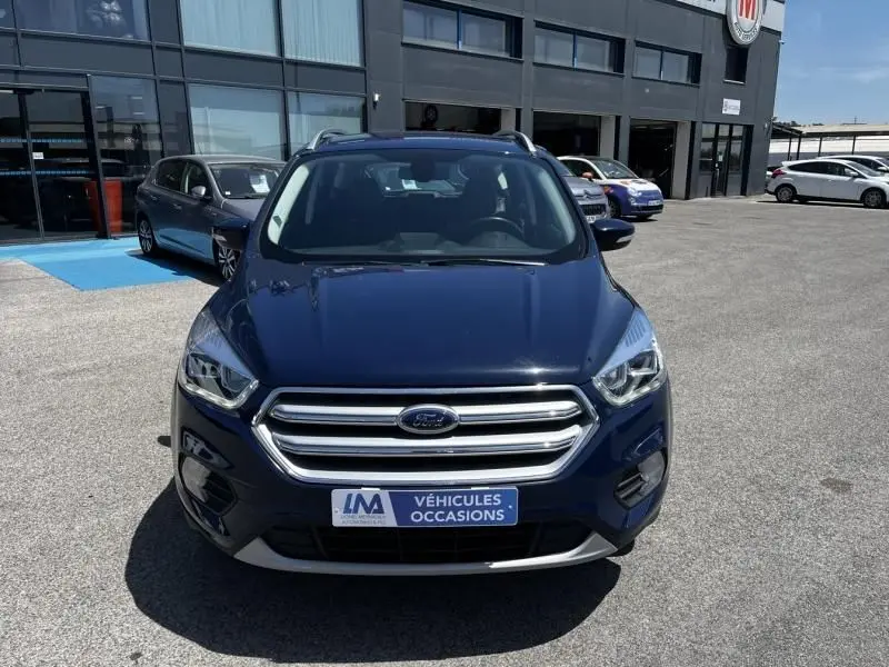 Vue frontale d'un Ford Kuga bleu foncé 2018 avec calandre chromée, stationné devant un bâtiment commercial.