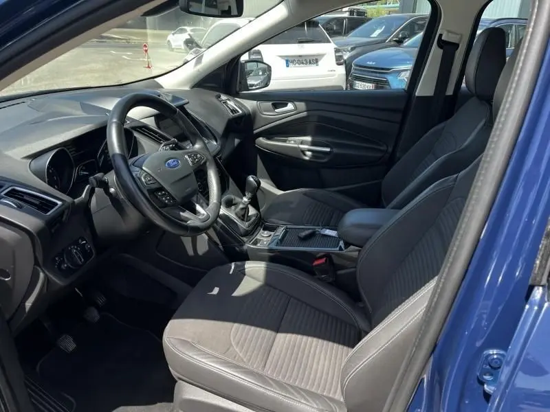 Vue intérieure côté conducteur du Ford Kuga bleu foncé 2018, montrant le volant, la boîte manuelle et les sièges en tissu gris.