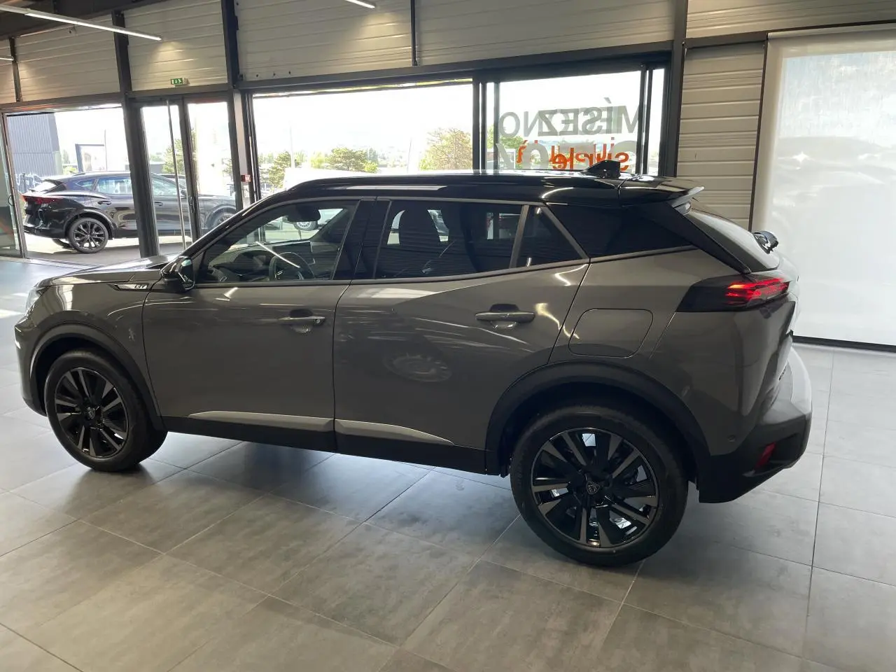 Peugeot 2008 gris en vue de profil côté gauche, avec jantes noires et toit noir contrastant, dans un showroom lumineux.