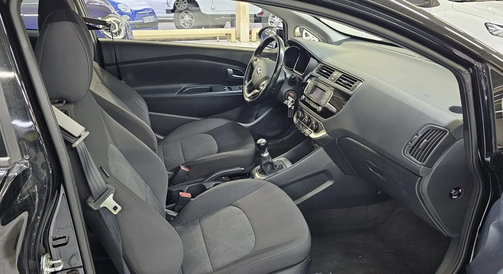 Intérieur noir de la KIA RIO III 3 portes, vue côté conducteur avec tableau de bord et boîte manuelle visibles.
