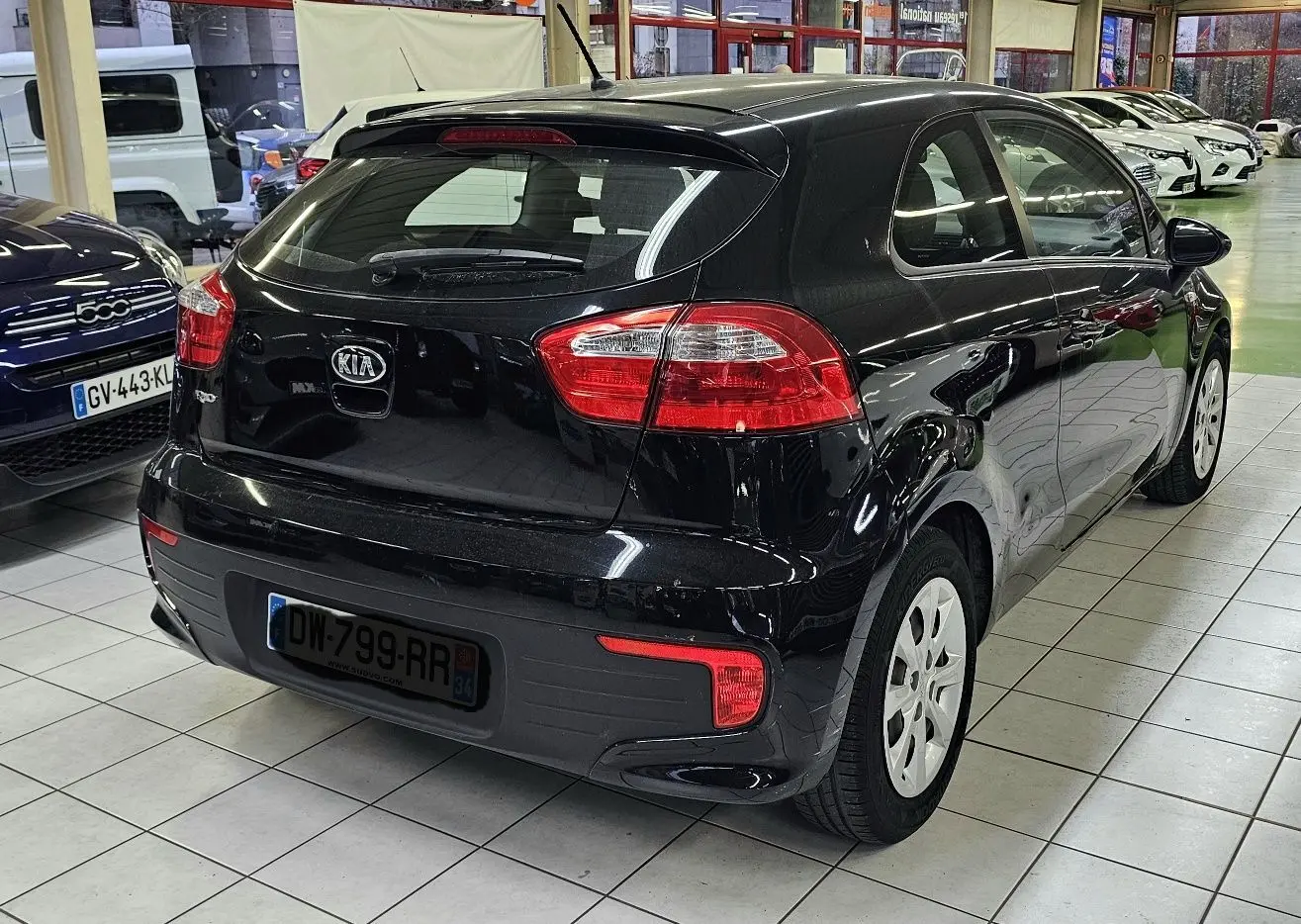 Vue 3/4 arrière droite d'une KIA RIO III noire 3 portes, avec feux arrière rouges et jantes blanches en showroom.