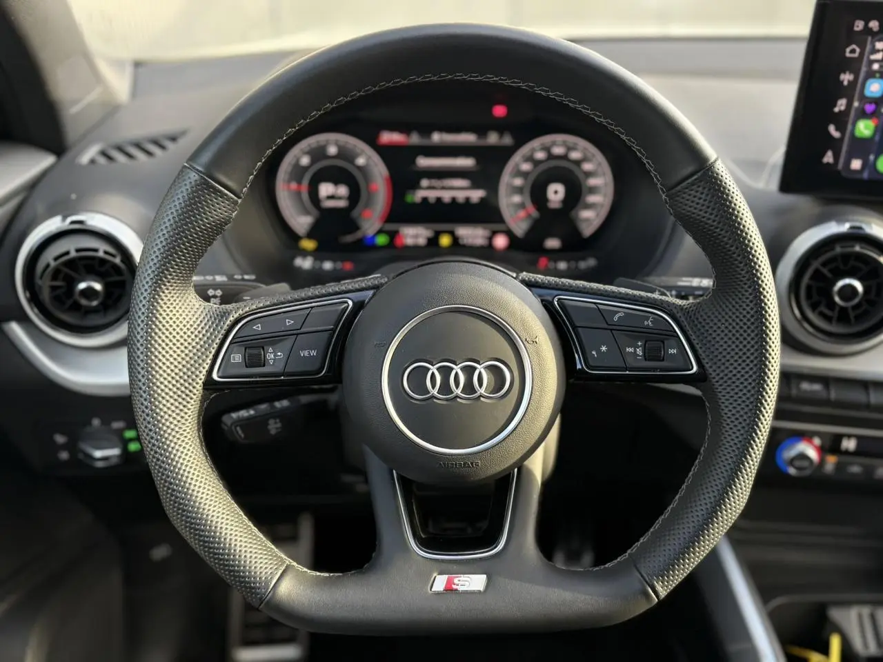 Vue rapprochée du volant cuir multifonction Audi S-Line avec commandes intégrées et tableau de bord numérique flou en arrière-plan.