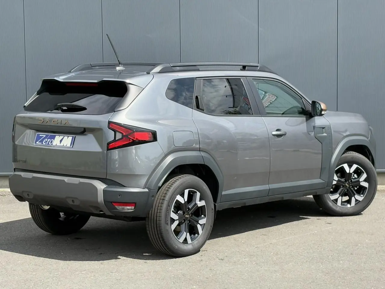Dacia Duster gris schiste en 3/4 arrière droit, avec jantes alliage noires et coques de rétroviseur brun cuivré.