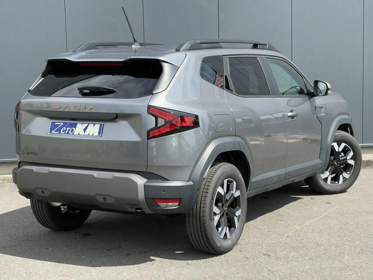 Vue 3/4 arrière droite du Dacia Duster 2025 gris schiste avec jantes alliage et barres de toit noires.