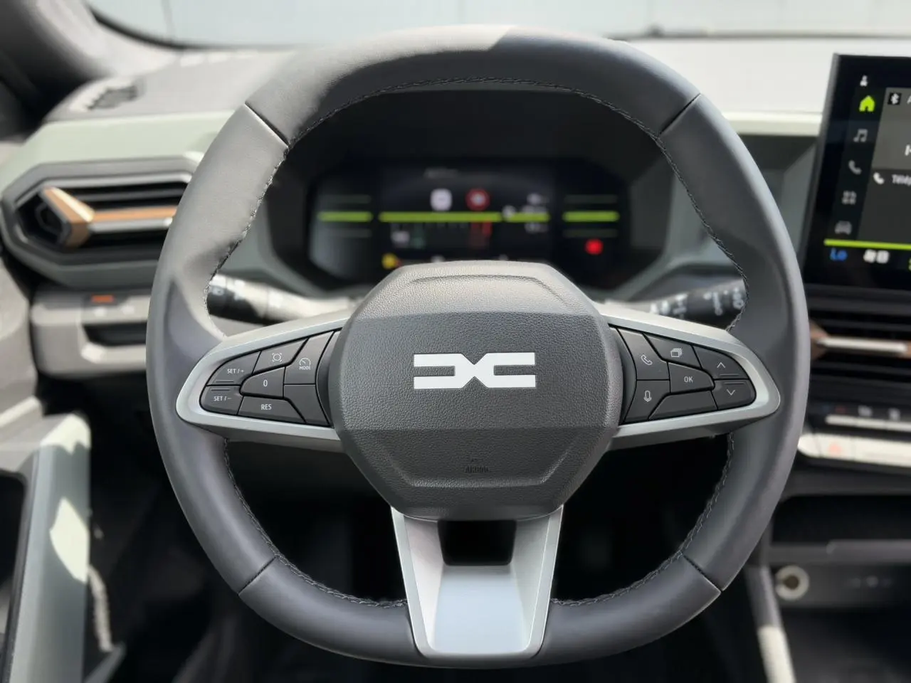 Volant multifonctions noir et gris du Dacia Duster 2025 avec tableau de bord digital et écran tactile visible en arrière-plan.