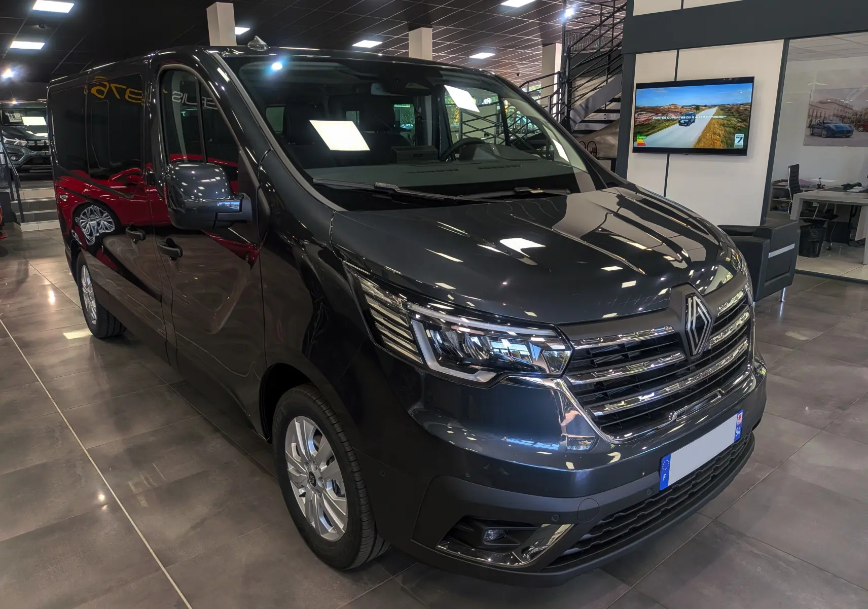 Renault Trafic Combi gris comète en 3/4 avant droit, intérieur showroom avec éclairage et reflet au sol.