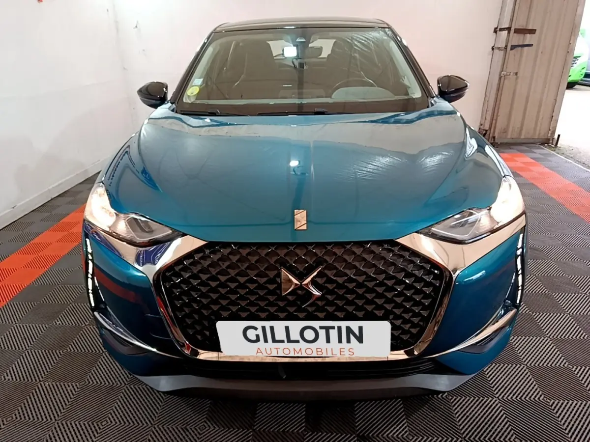 Vue frontale d'une DS3 Crossback bleu métallisé avec calandre noire à motifs géométriques et phares allumés.