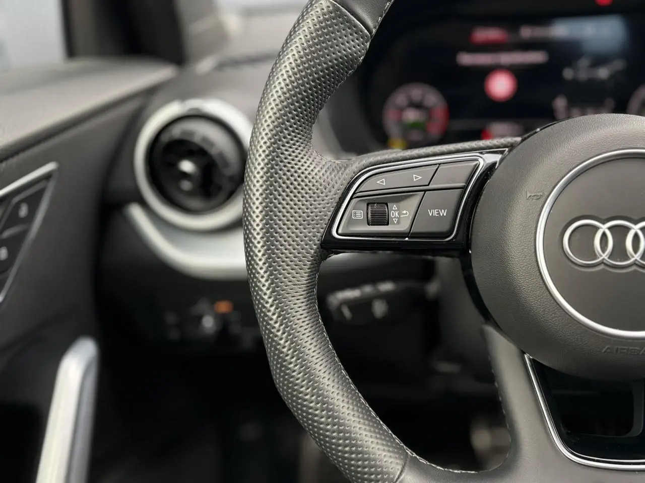 Gros plan sur le volant en cuir perforé de l'Audi Q2 35 TDI S-Line, avec commandes multifonctions et aérateur rond à gauche.