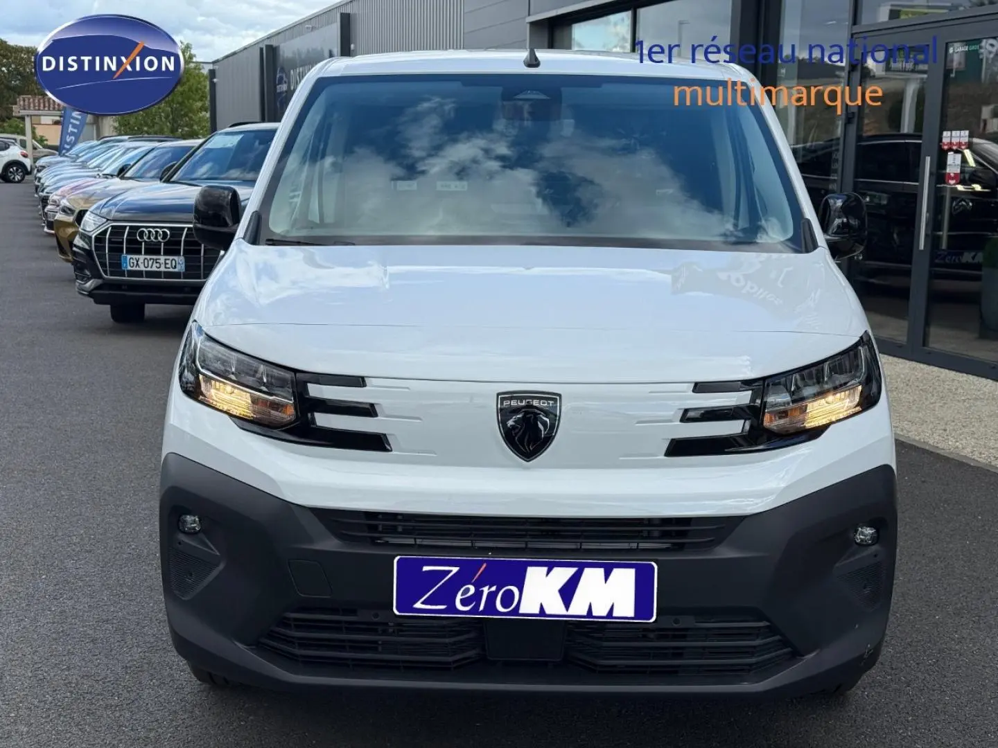 Vue frontale d'un Peugeot Partner blanc Icy 2026 avec feux allumés et pare-chocs noir mat visible en concession.