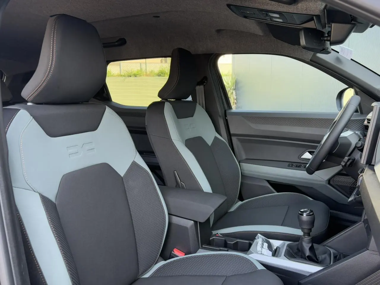 Intérieur avant droit du Dacia Duster 2025, sièges bi-ton gris clair et noir avec volant et levier de vitesse visibles.