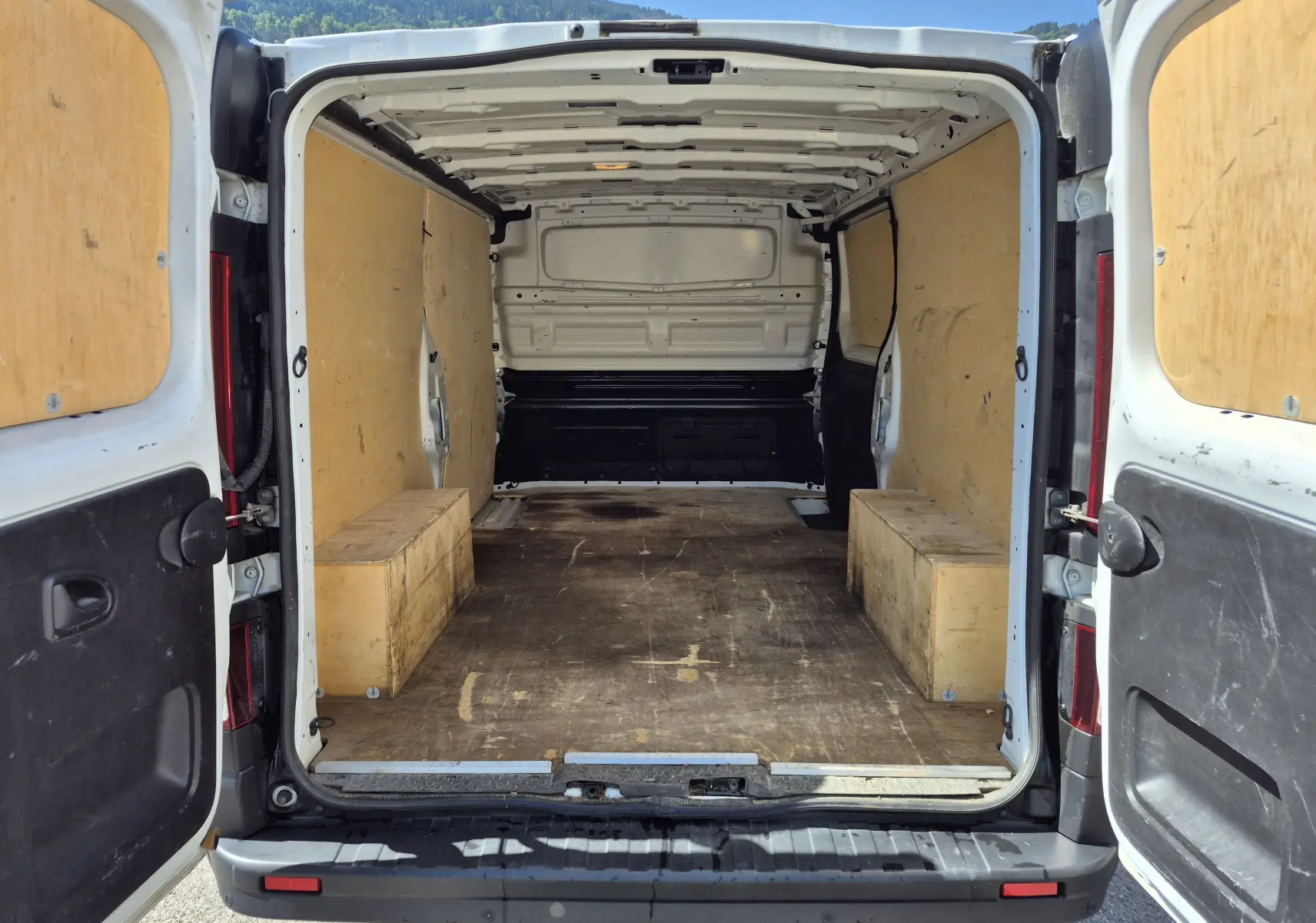 Vue arrière du Nissan Primastar blanc avec portes ouvertes, montrant l'intérieur vide et aménagé en bois du fourgon.
