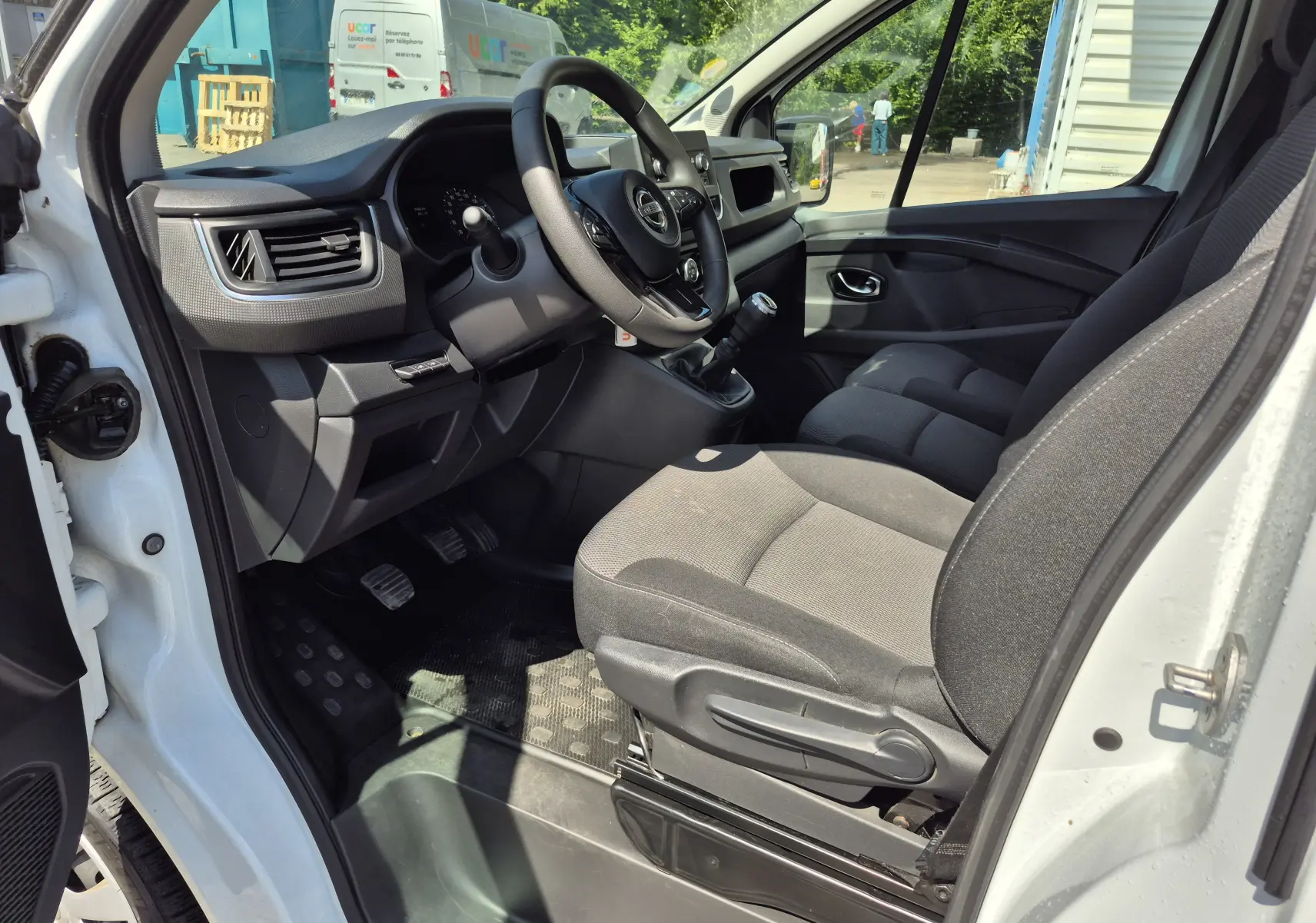 Vue intérieure côté conducteur du Nissan Primastar blanc, montrant le tableau de bord, volant multifonction et sièges en tissu gris.