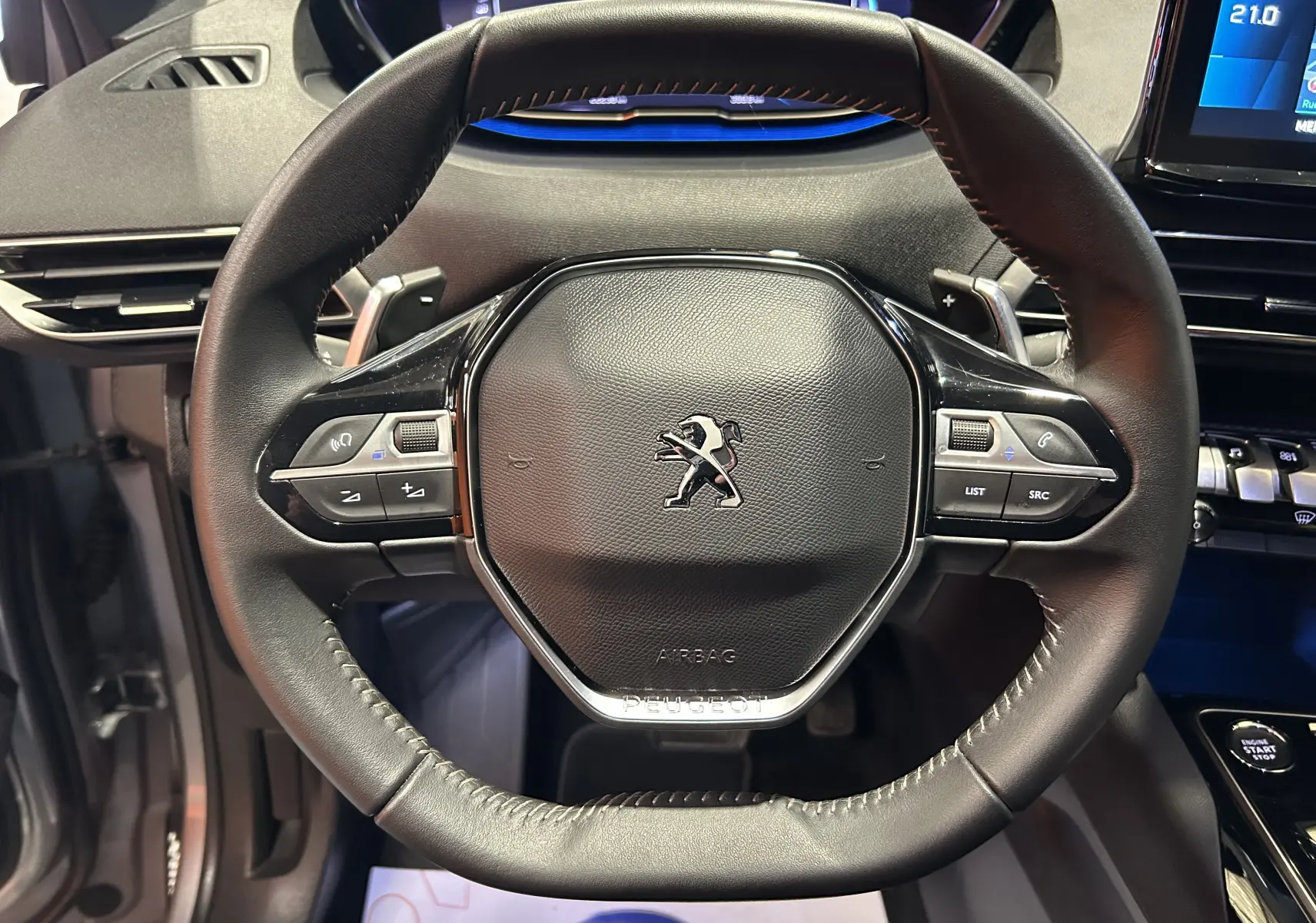 Gros plan sur le volant cuir noir du Peugeot 3008 Hybrid 2024, avec commandes intégrées et tableau de bord numérique en arrière-plan.