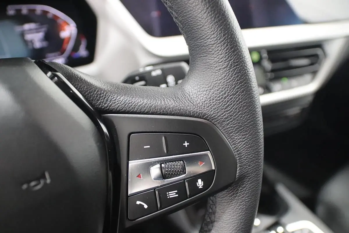 Gros plan sur les commandes du volant en cuir noir de la BMW Série 1 116d 2023 avec affichage numérique en arrière-plan.