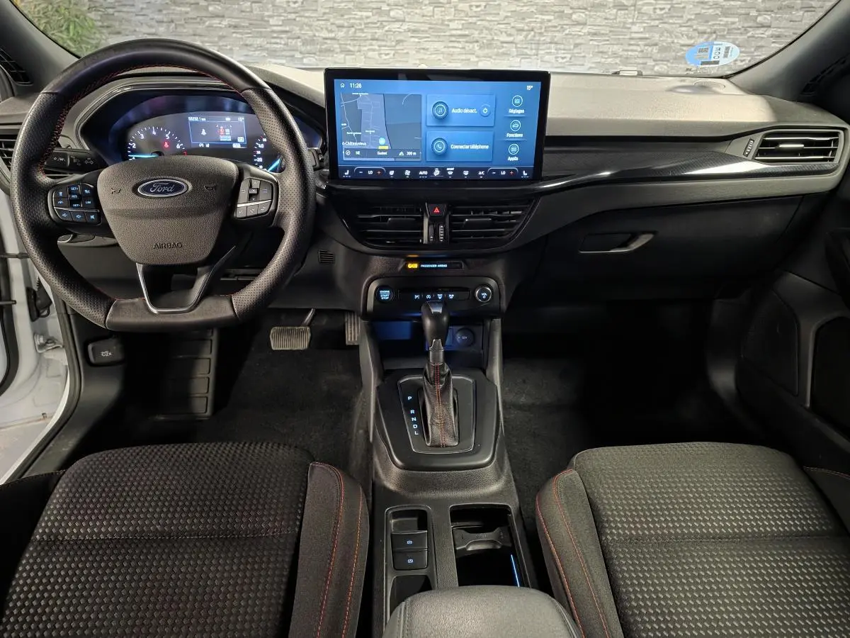Vue intérieure avant de la Ford Focus 2024 blanche, tableau de bord noir avec écran tactile central et volant ST Line à surpiqûres rouges.