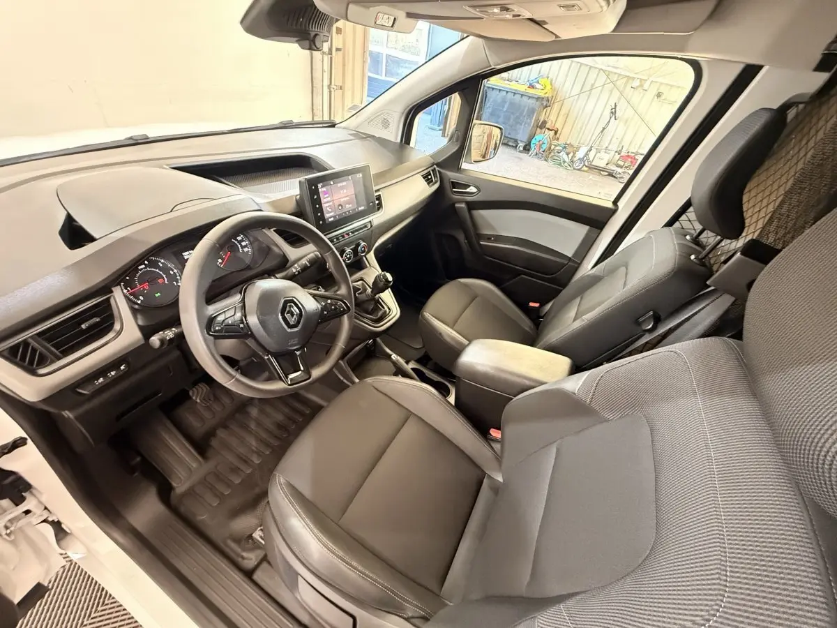 Vue intérieure côté conducteur du Renault Kangoo blanc 2022, montrant le tableau de bord et les sièges avant en tissu gris.