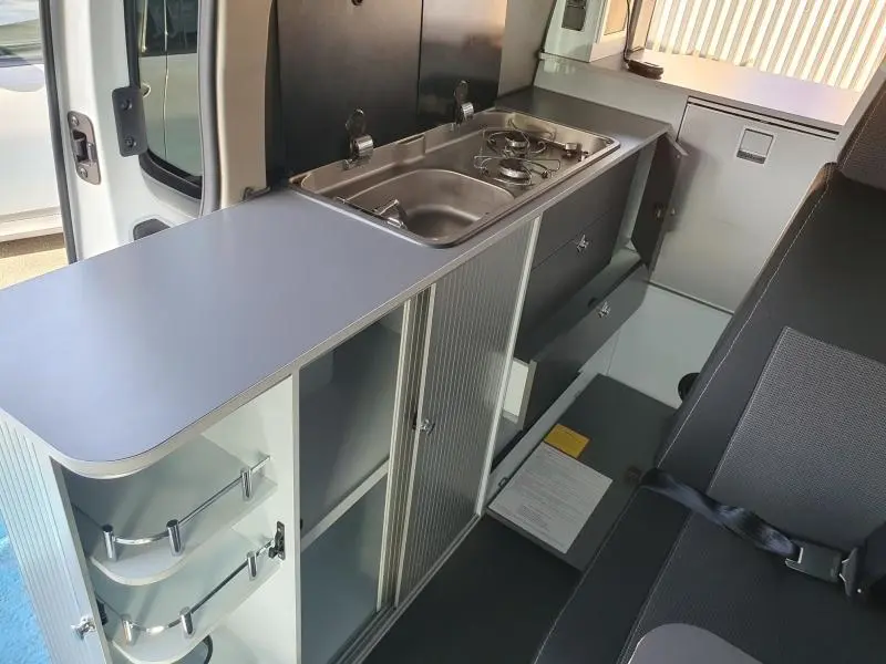 Vue intérieure côté droit du Renault Trafic Fourgon blanc glacier, montrant la kitchenette avec évier et plaque de cuisson intégrés.