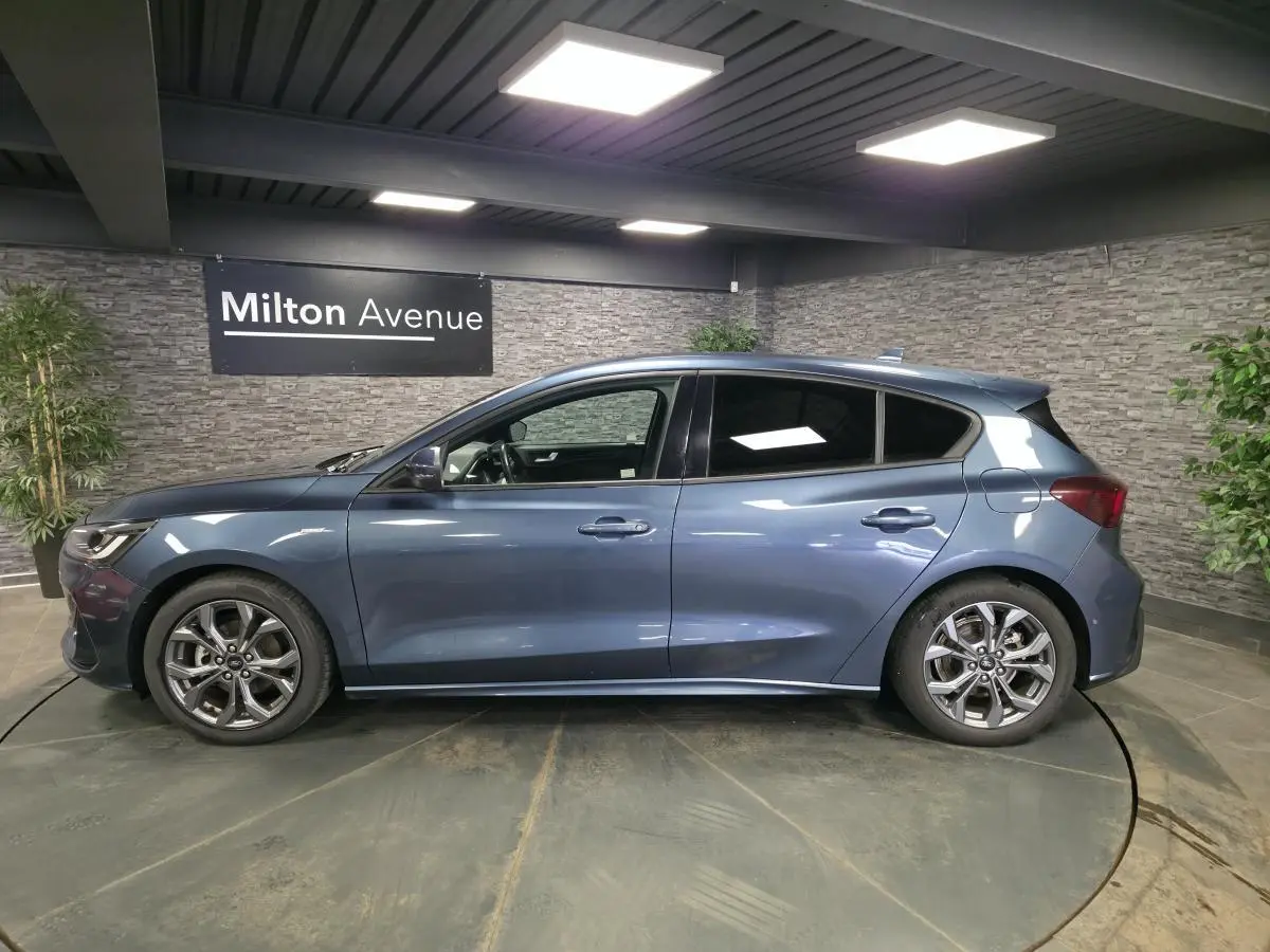 Profil gauche d'une Ford Focus 2024 bleu azur avec jantes alliage 17'' et vitres arrière surteintées dans un showroom.