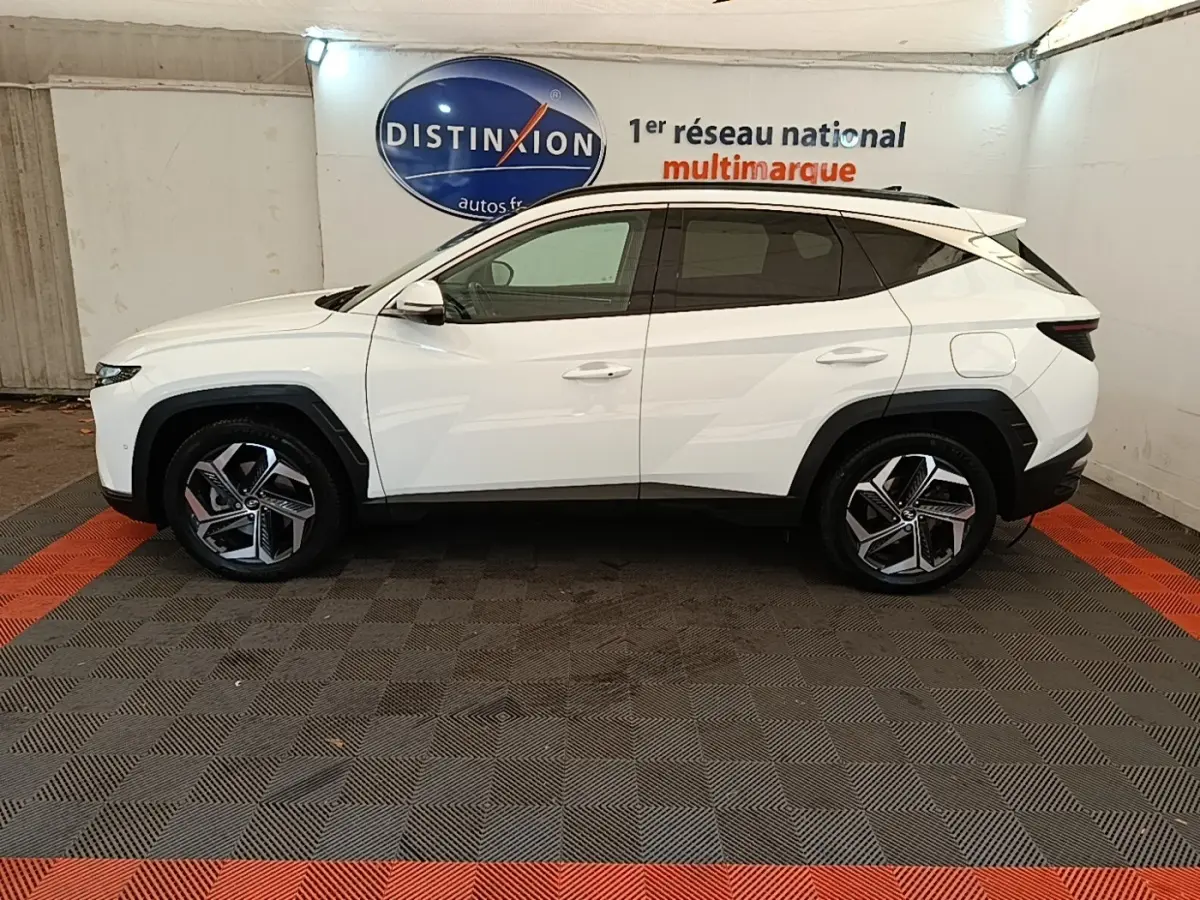 Vue latérale gauche d'un Hyundai Tucson blanc 2022 hybride, avec jantes alliage et toit noir contrasté en intérieur showroom.
