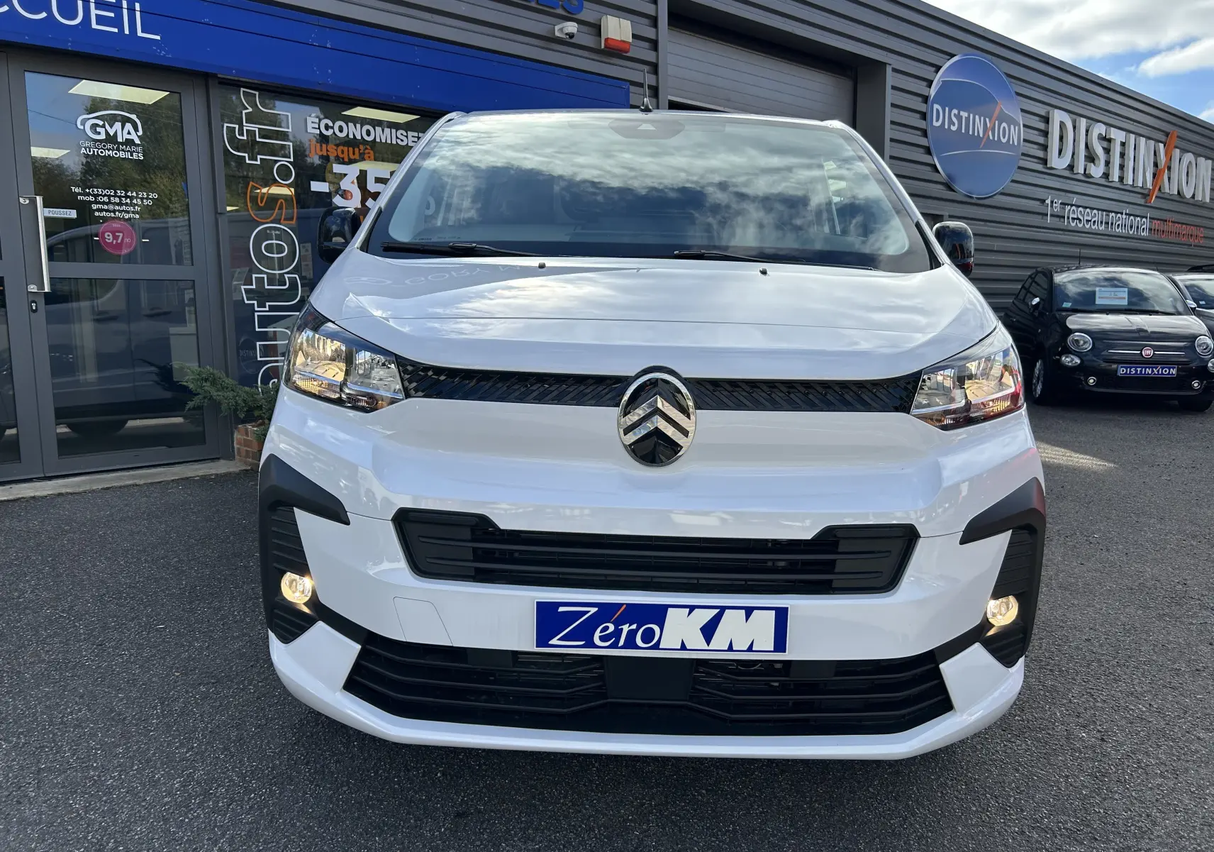 Vue avant d’un Citroën Jumpy blanc 2025 avec calandre noire et phares allumés, stationné devant un bâtiment commercial.