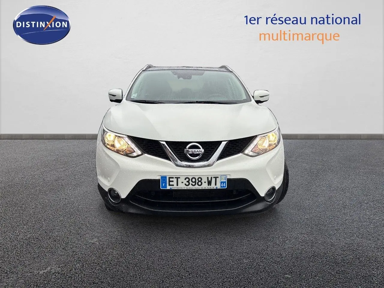 Vue frontale d'un Nissan Qashqai blanc glacier de 2017 avec phares allumés sur sol gris.