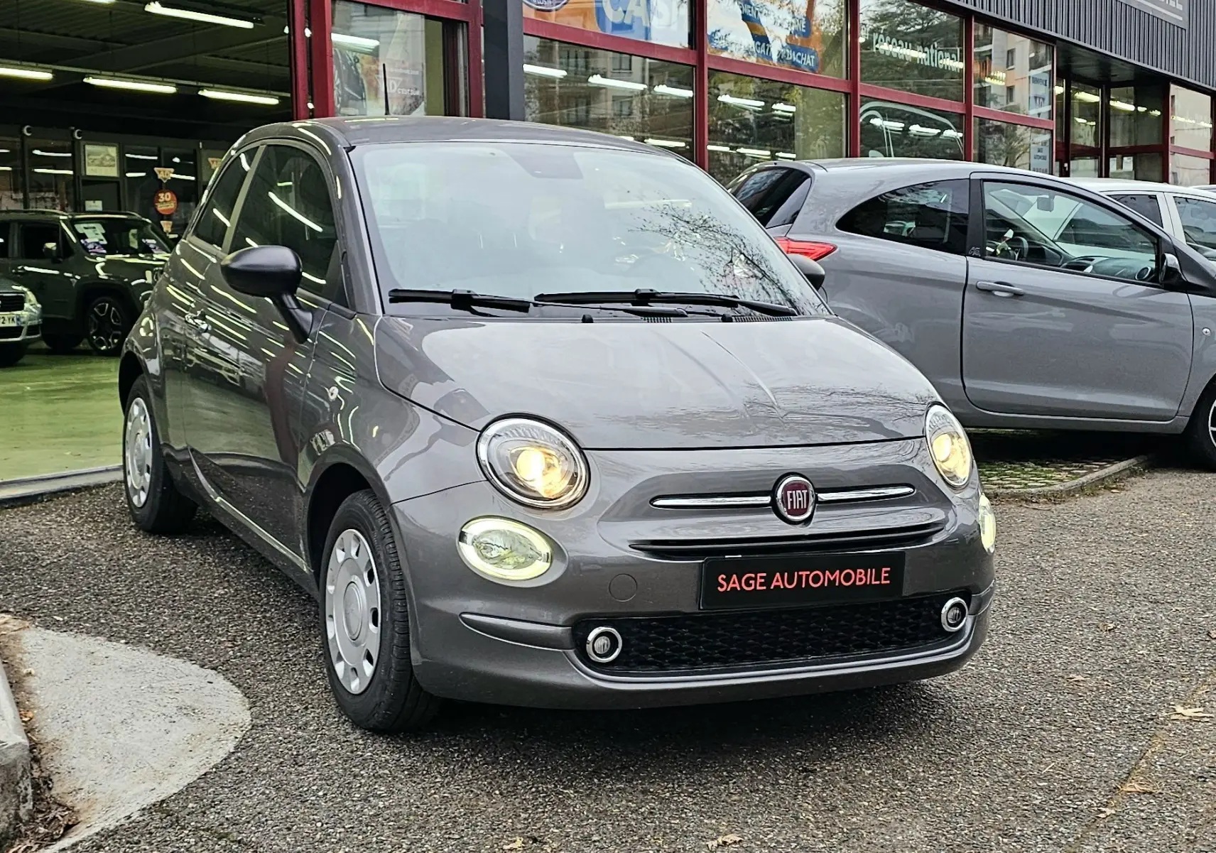 Fiat 500 gris vue 3/4 avant droit, phares allumés, stationnée devant un garage Sage Automobile.