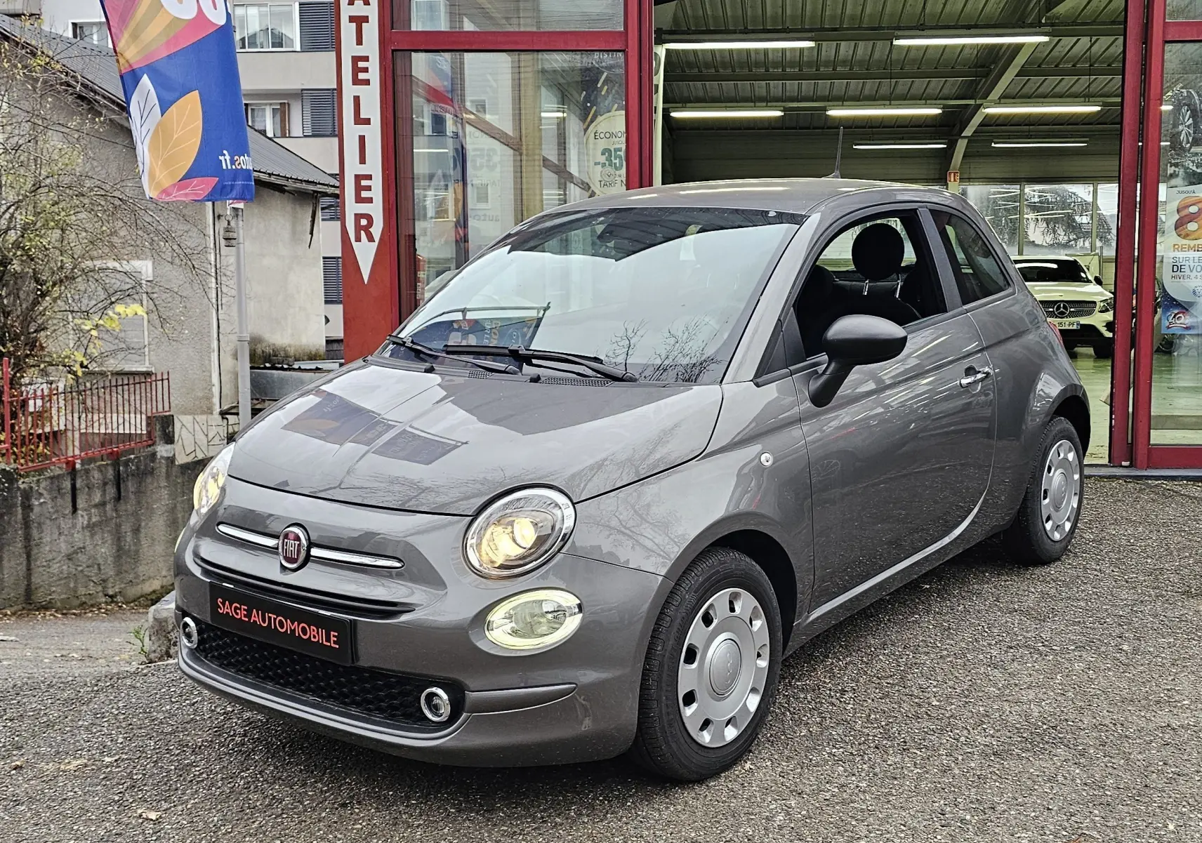 FIAT 500 gris vue 3/4 avant droit avec phares allumés, garée devant un atelier automobile.