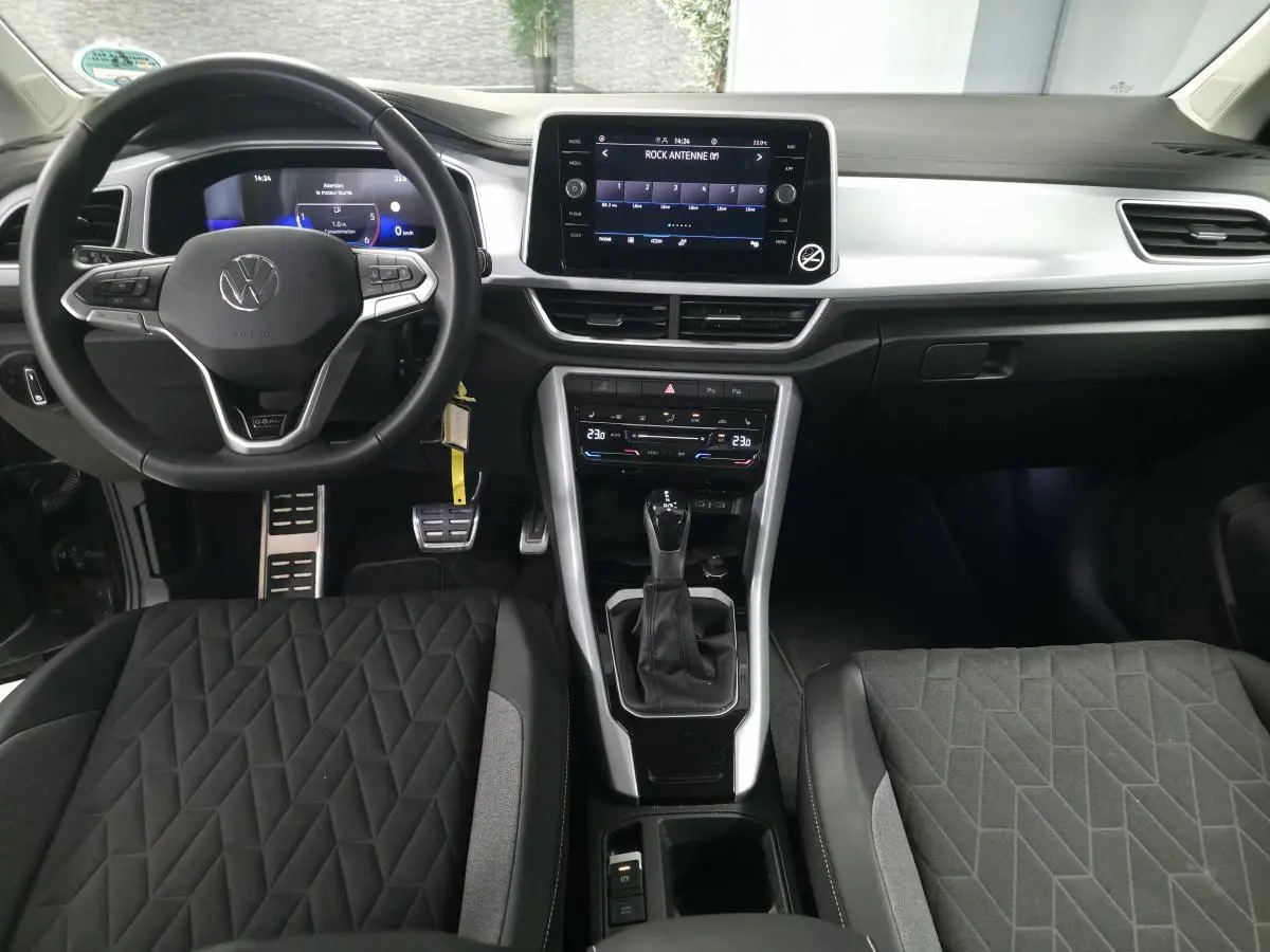 Vue intérieure avant du Volkswagen T-Roc 2025 noir, avec volant multifonction, écran tactile central et sièges tissu noir à motifs géométriques.