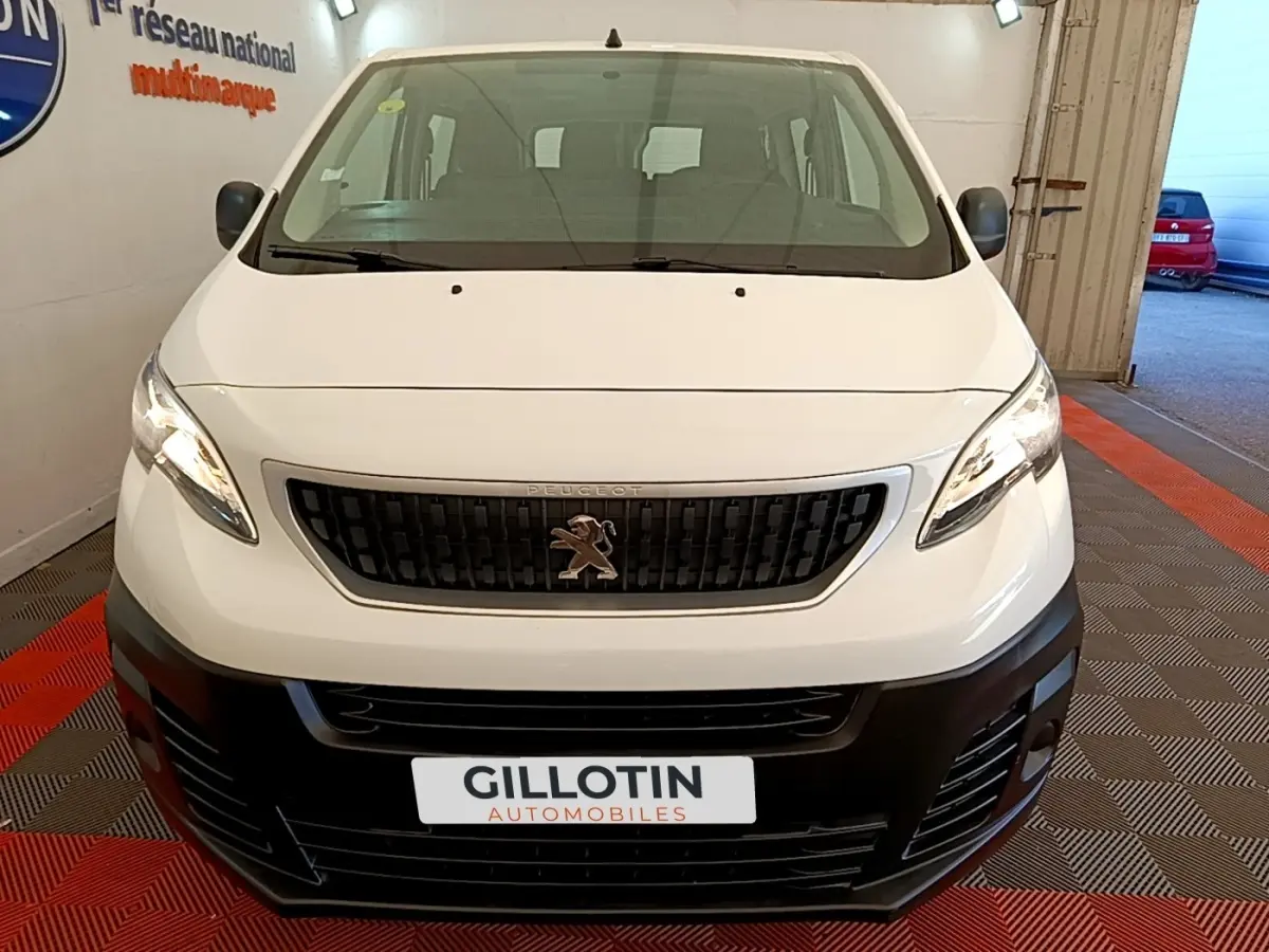 Vue avant d’un Peugeot Traveller blanc de 2018 avec calandre noire et logo lion visible, stationné en intérieur.