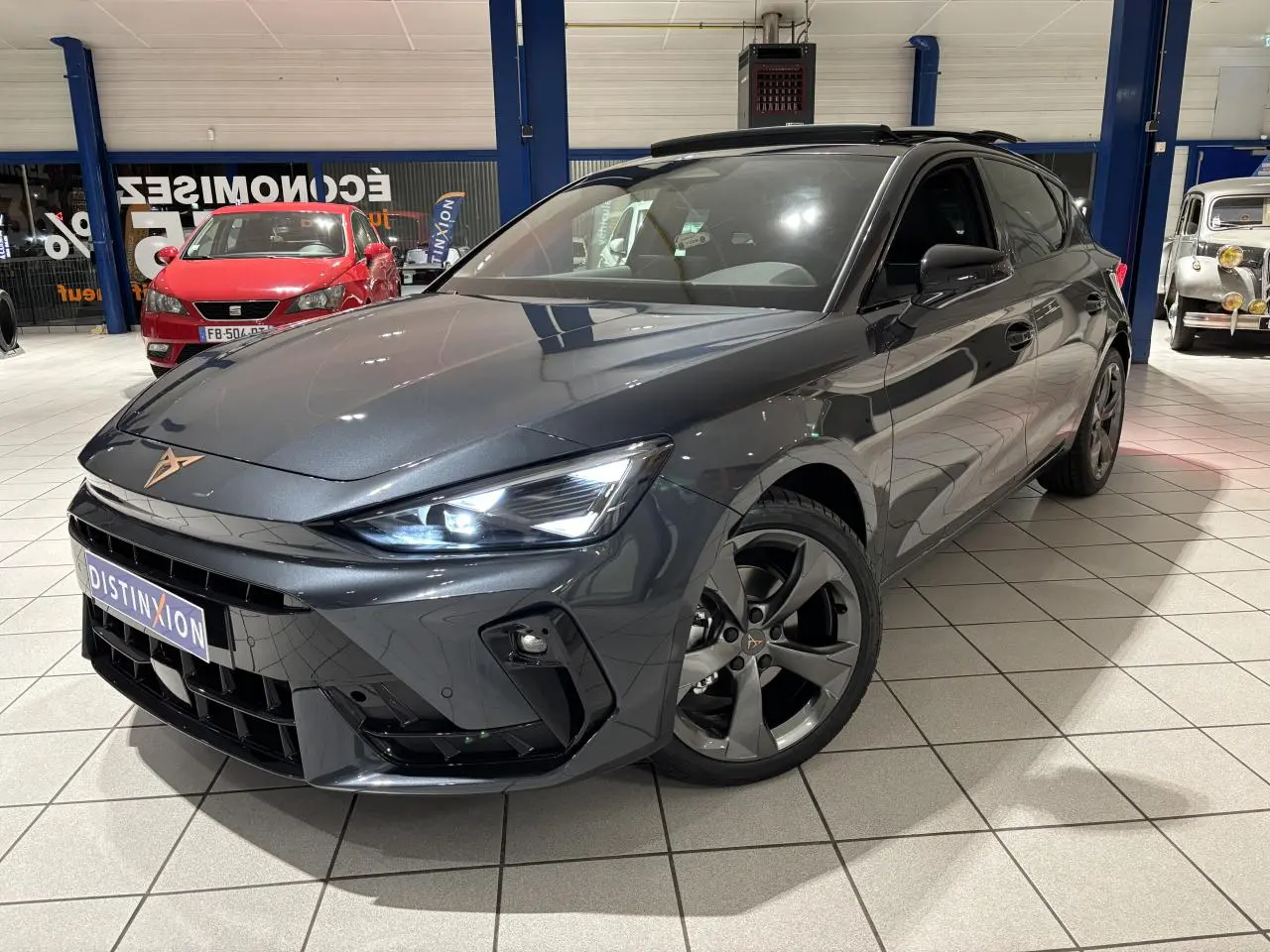 Vue 3/4 avant d'une CUPRA Leon gris Graphène avec jantes anthracite et signature lumineuse Tri-LED visible en intérieur showroom.