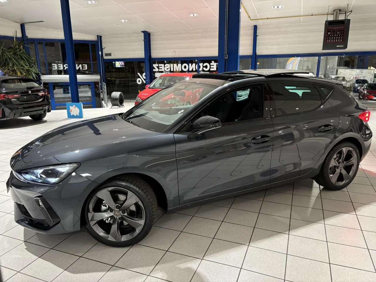 Vue de profil côté gauche d’une CUPRA Leon gris Graphène avec jantes anthracite en showroom lumineux.