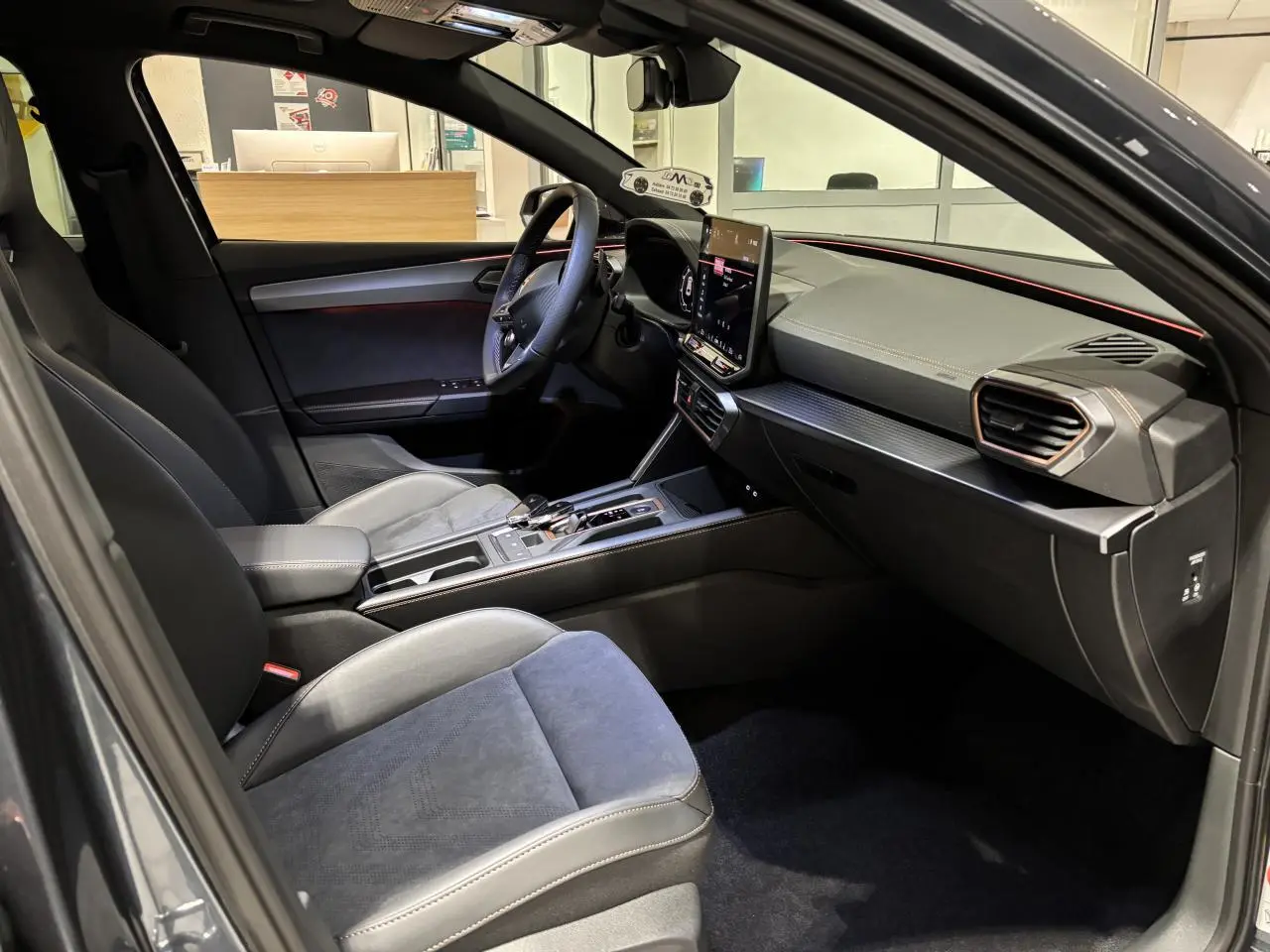 Vue intérieure côté conducteur de la CUPRA Leon 2026 gris Graphène avec sièges suédine noirs et tableau de bord moderne.