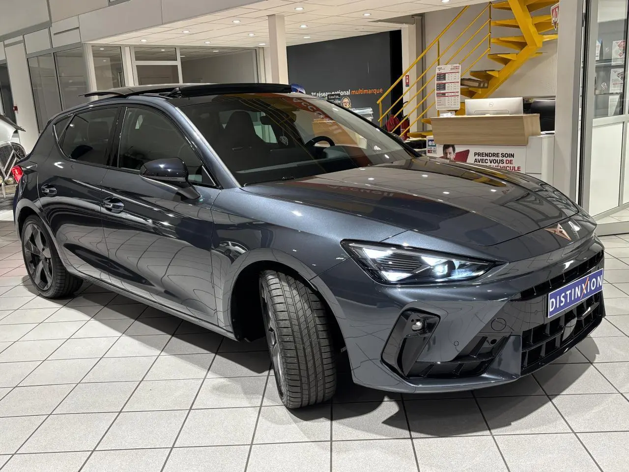 Vue 3/4 avant droite d'une CUPRA Leon gris Graphène avec phares LED allumés et jantes anthracite en showroom.