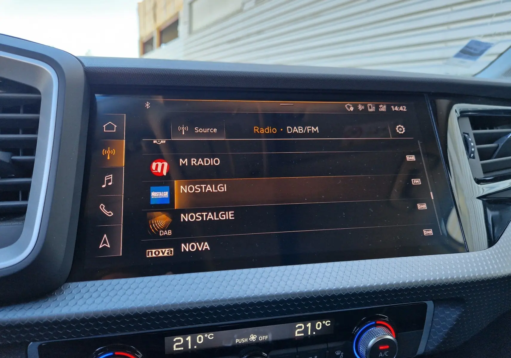 Vue rapprochée de l’écran multimédia et de la console de climatisation de l’Audi A1 Sportback blanc glacier, affichant les radios DAB/FM.