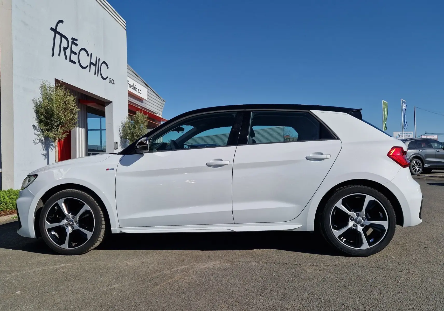 Profil droit d'une Audi A1 Sportback blanc glacier avec toit noir et jantes alliage distinctives, stationnée en extérieur.