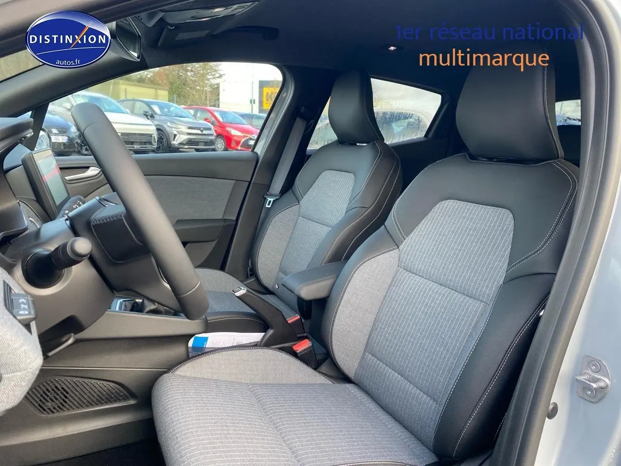 Vue intérieure côté conducteur de la Renault Clio V gris rafale, montrant les sièges tissu et cuir avec accoudoir central.