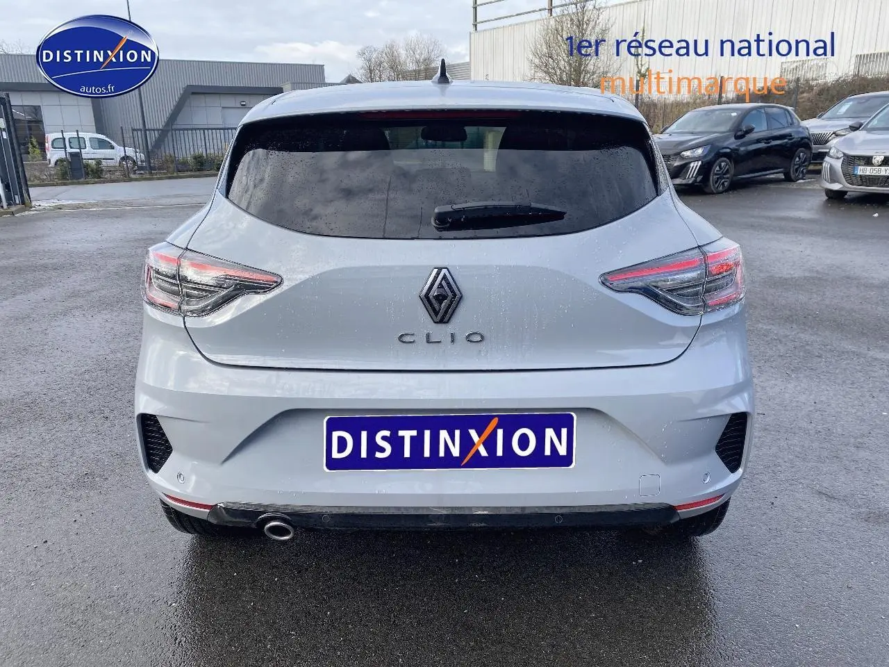 Vue arrière d'une Renault Clio V gris rafale avec feux LED et sortie d'échappement chromée sur parking humide.