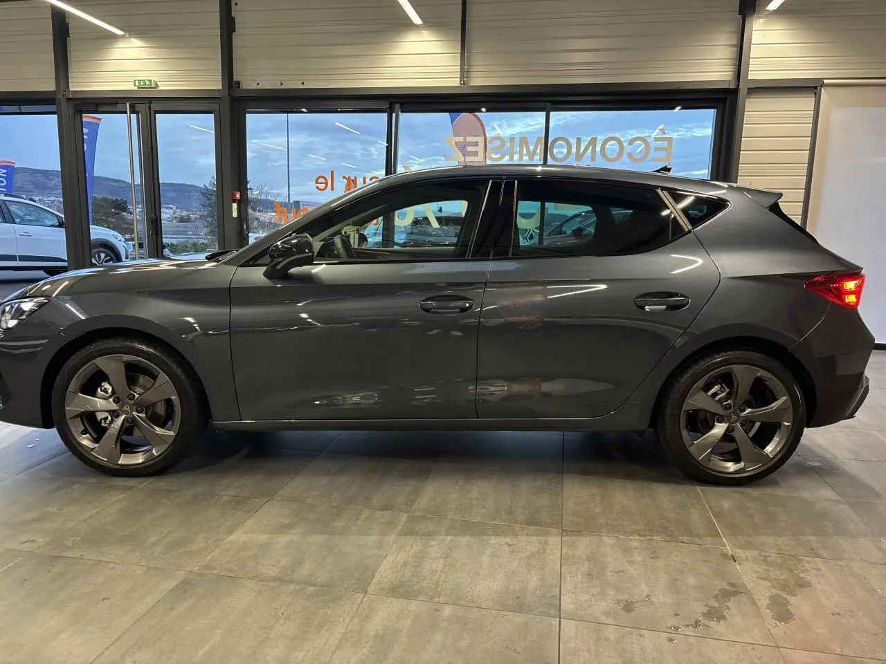 Vue de profil droit d'une CUPRA Leon 2.0 TDI gris magnetic avec jantes alliage 18 pouces en intérieur showroom.