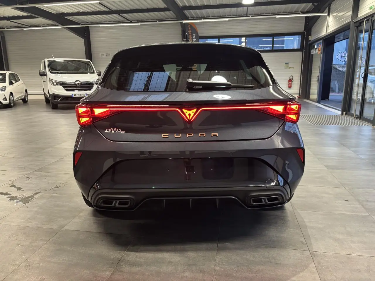 Vue arrière d'une CUPRA Leon gris magnetic avec feux arrière Tri-LED et double sortie d'échappement noire en intérieur showroom.