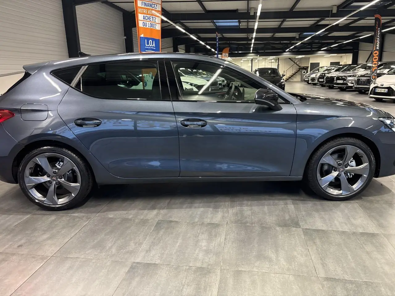 Vue de profil côté gauche d'une CUPRA Leon gris magnetic avec jantes alliage 18 pouces dans un showroom.