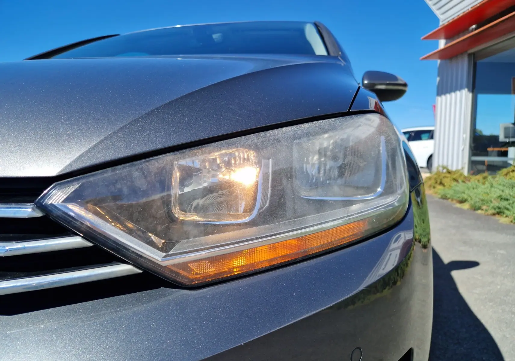 Gros plan sur le phare avant droit allumé d'une Volkswagen Golf Sportsvan gris Indium Metal sous un ciel bleu.