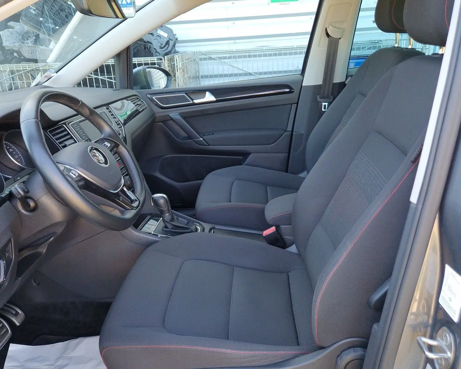 Vue intérieure côté conducteur de la Volkswagen Golf Sportsvan gris Indium, sièges tissu noir avec surpiqûres rouges.
