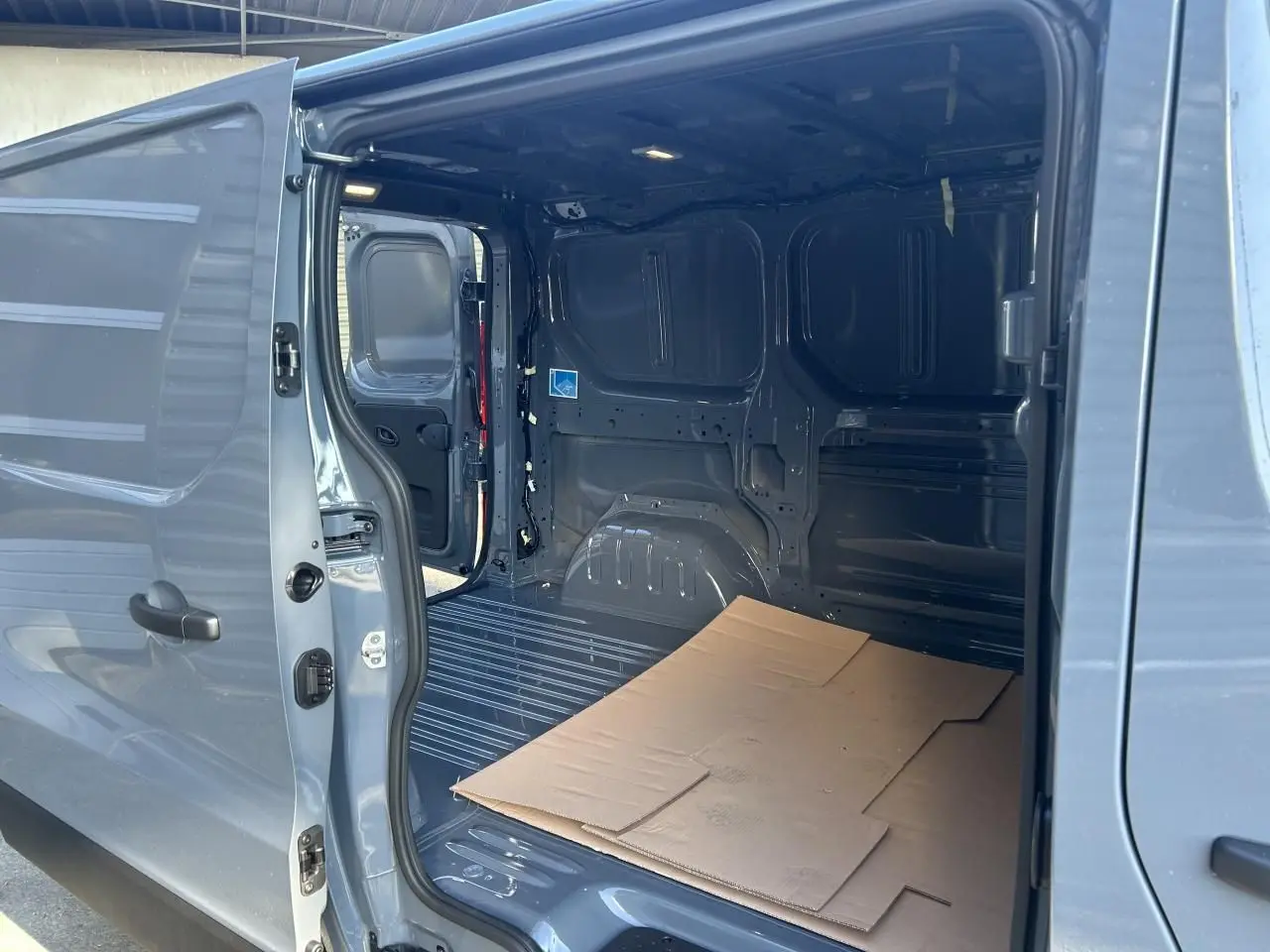 Vue intérieure du Renault Trafic Fourgon gris urbain, porte latérale ouverte montrant l'espace de chargement vide avec carton au sol.
